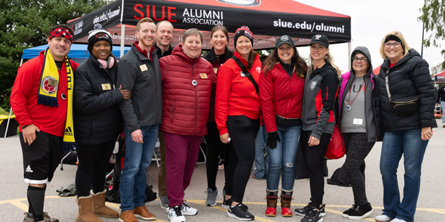 A group shot of five adults, and one baby, wearing SIUE gear next to a sports field