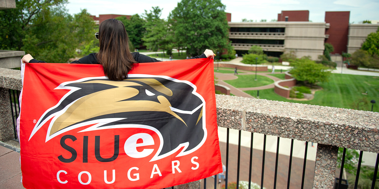 Female student holding a SIUE Cougar flag while standing on the second floor balcony of Rendleman Hall.