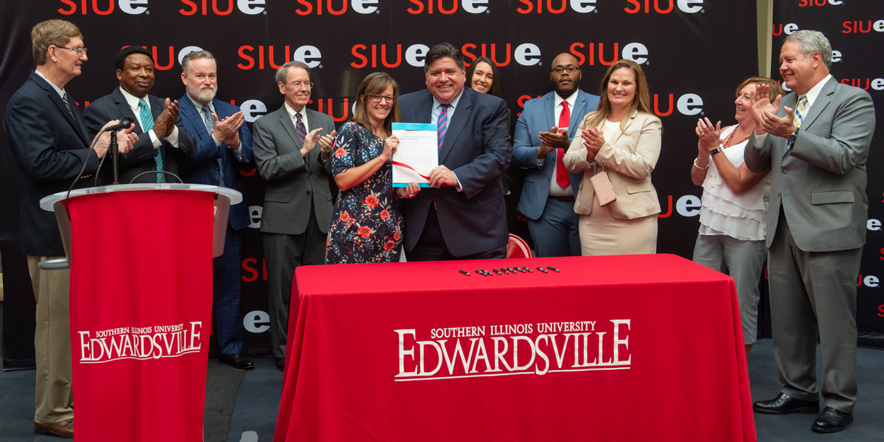 Illinois Governor J.B. Pritzker celebrates the signing of House Bill 2239 during a visit to SIUE’s campus. He stands alongside Rep. Katie Stuart and is surrounded by members of the SIU Board of Trustees and additional contributing legislators.