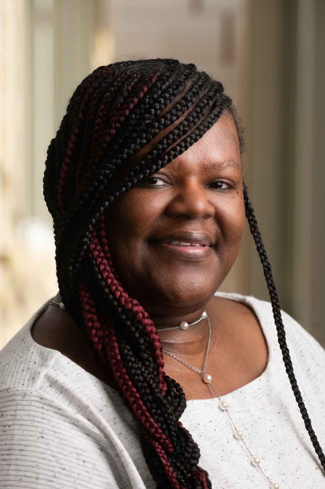 A snapshot of Calandra Mitchell, a melanated, African American woman with braided hair, a single strand pearl accented necklace adorned over a white, black-speckled dress. Calandra is smiling.