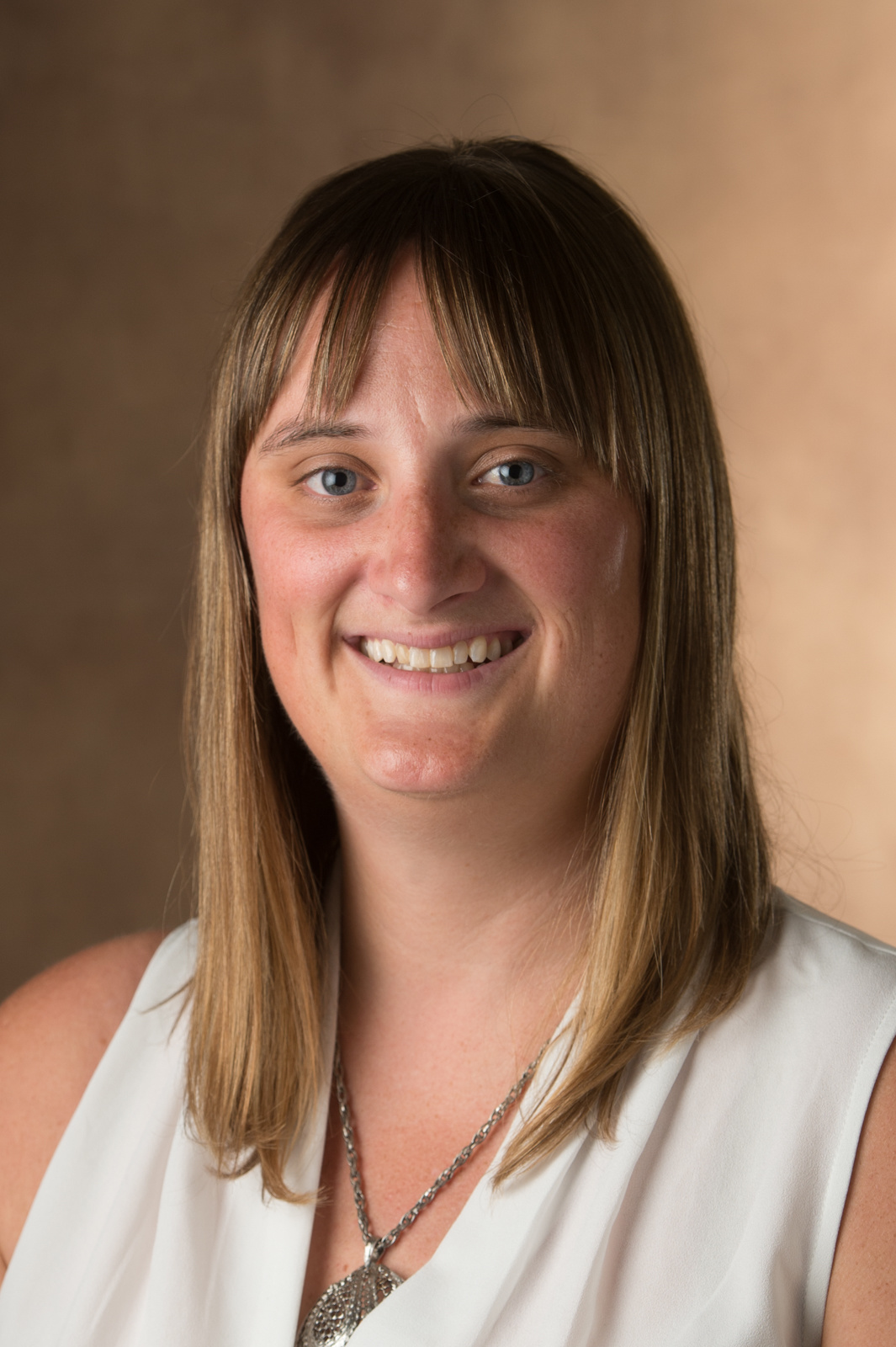 Portrait of Lindsay Ross-Stewart, she has medium-length straight dark blonde hair, bangs, a smile, a silver necklace and a white sleeveless blouse. 