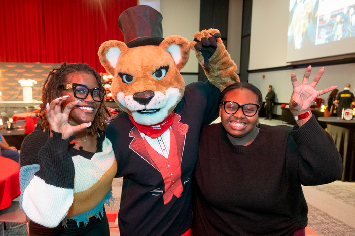 Two students on either side of Eddie the Cougar who is wearing a tuxedo and top hat. The students are making claws with their hands in a fun pose to mimic the Cougar