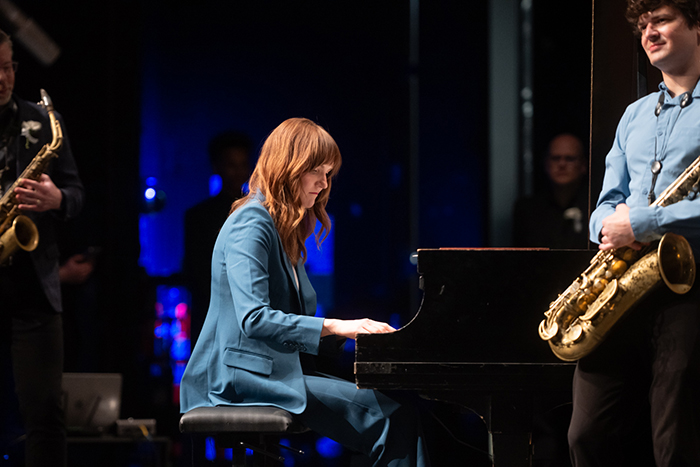 Baldus gracefully seated at the piano. She is wearing a blue suit, her eyes focused on the keys and her face serene