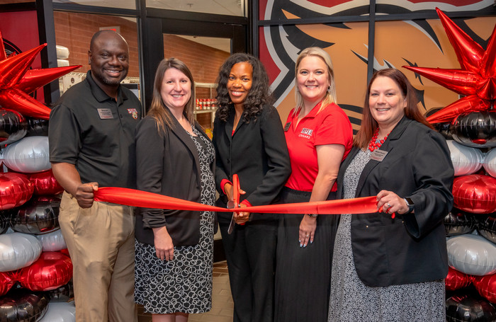 Photo of Rony Die, Andrea Keller, Lealia Williams, Mindy Dilley, and Miriam Roccia holding a red ribbon. Williams is holding a large scissors and poised to make the cermonial cut. They are in front of the Cougar Cupboard's new location and mylar balloon pillars surround them on both sides