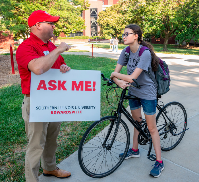 A staff member holding an "Ask Me!" sign with the University wordmark stands in the quad and assists a girl on a black bicycle