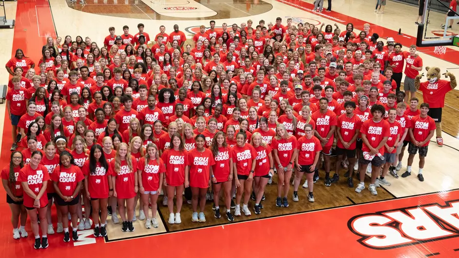 The collective group of SIUE's student athletes gathered in an overhead shot in the Vadalabene Center First Community Arena. The student-athletes area all wearing red branded SIUE shirts with the Cougar logo