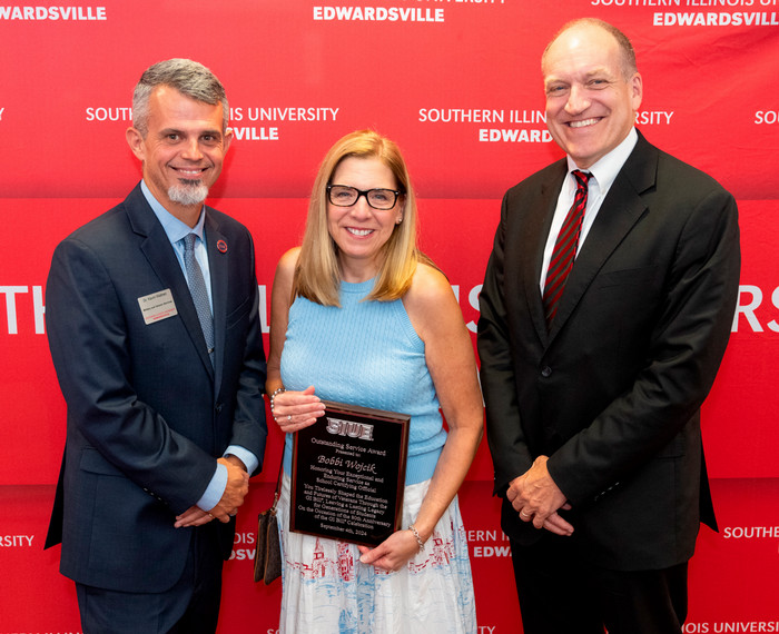 Group photo in front of a red SIUE-branded background of Kevin Wathen, Bobbi Wojcik, and Dan Mahony. Bobbi is holding an SIUE plaque with the Outstanding Service Award and her name engraved on it.