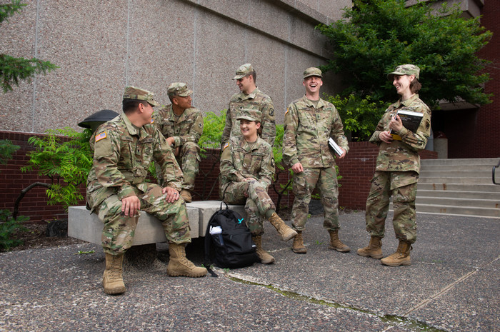 A Group of six military students wearing army fatigues, laughing around a meditation chair outside Peck Hall. They have bookbags and are holding school supplies