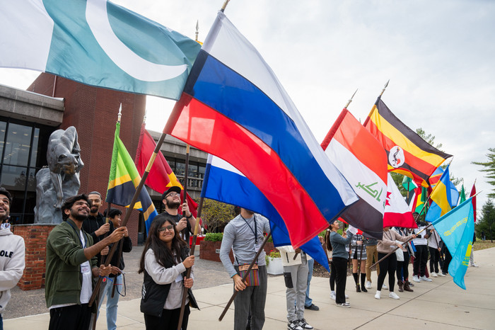 A group of international students standing in a line waving full sized flags of their home nation on a cloudy day outside the MUC and by the Cougar Statue